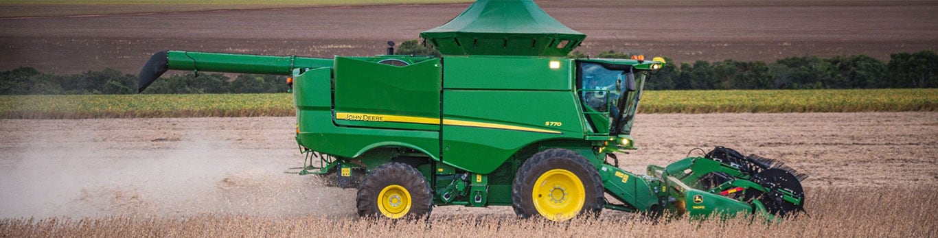 Una cosechadora John Deere 5770 trabajando en un campo, con cosechas doradas y un cielo despejado al fondo, con el polvo levantándose de su paso.
