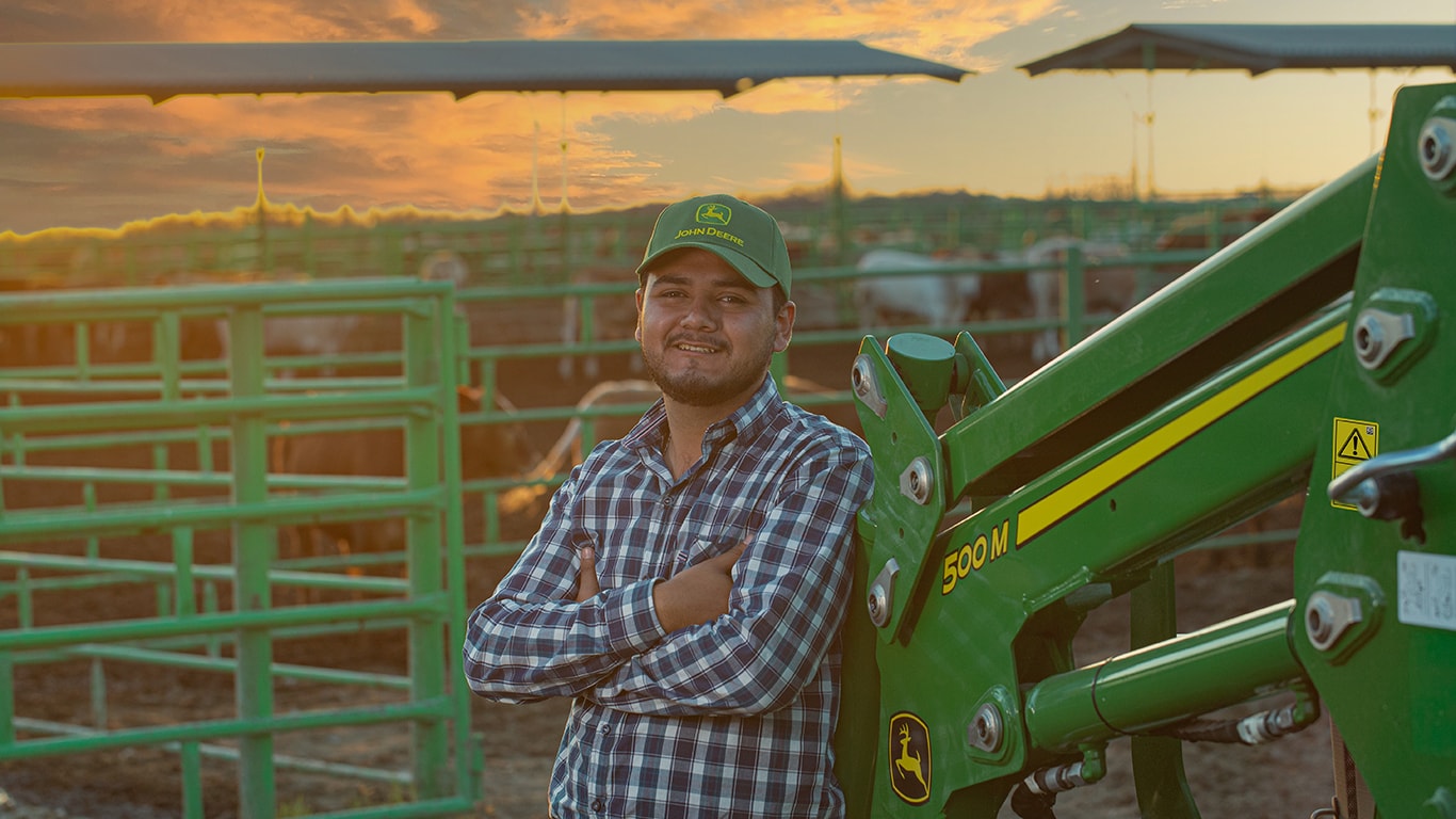 Jóven en camisa de cuadros y gorra John Deere, recargado en cargador frontal 500M con los brazos cruzados. De fondo está el atardecer y ganado en su establo.